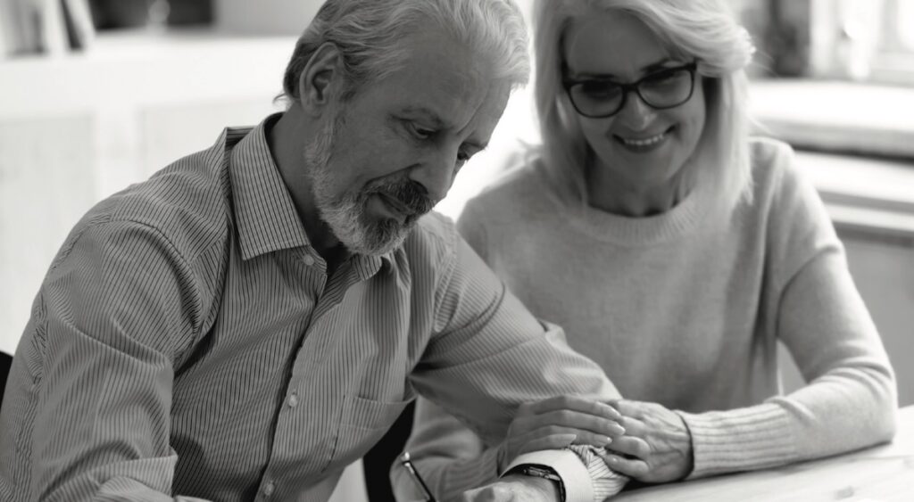 An older man signs a paper while his wife holds his arm with encouragement.