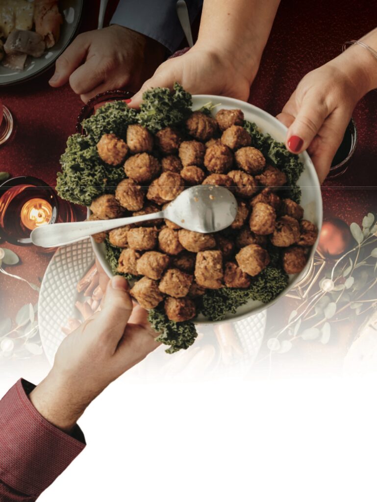 A round serving dish of meatballs with kale garnish and a silver serving spoon being passed between people's hands.