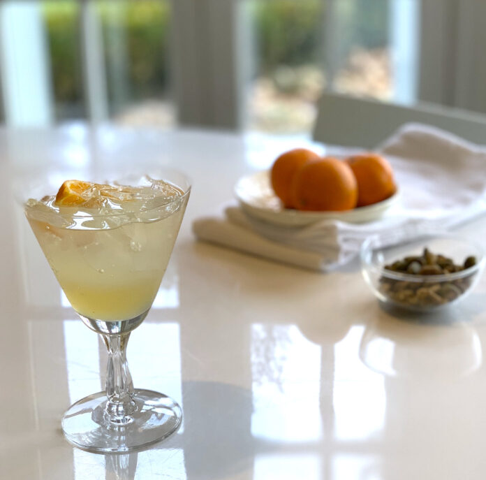 Tangy Lemon Ginger Shrub A lemon ginger shrub cocktail sits in a triangle glass, the liquid is light yellow in color. Behind the drink are three oranges on a cloth napkin.