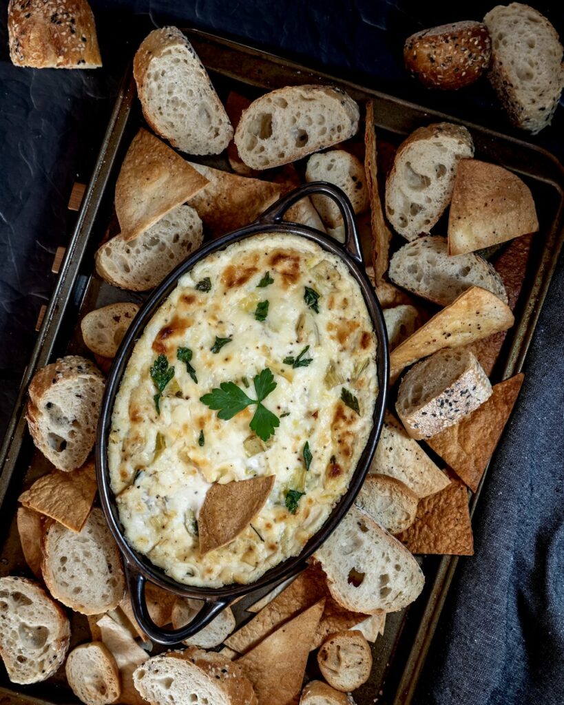 Top down view of a cookie sheet filled with various pieces of bread and pita triangles and a warm artichoke dip in the middle.