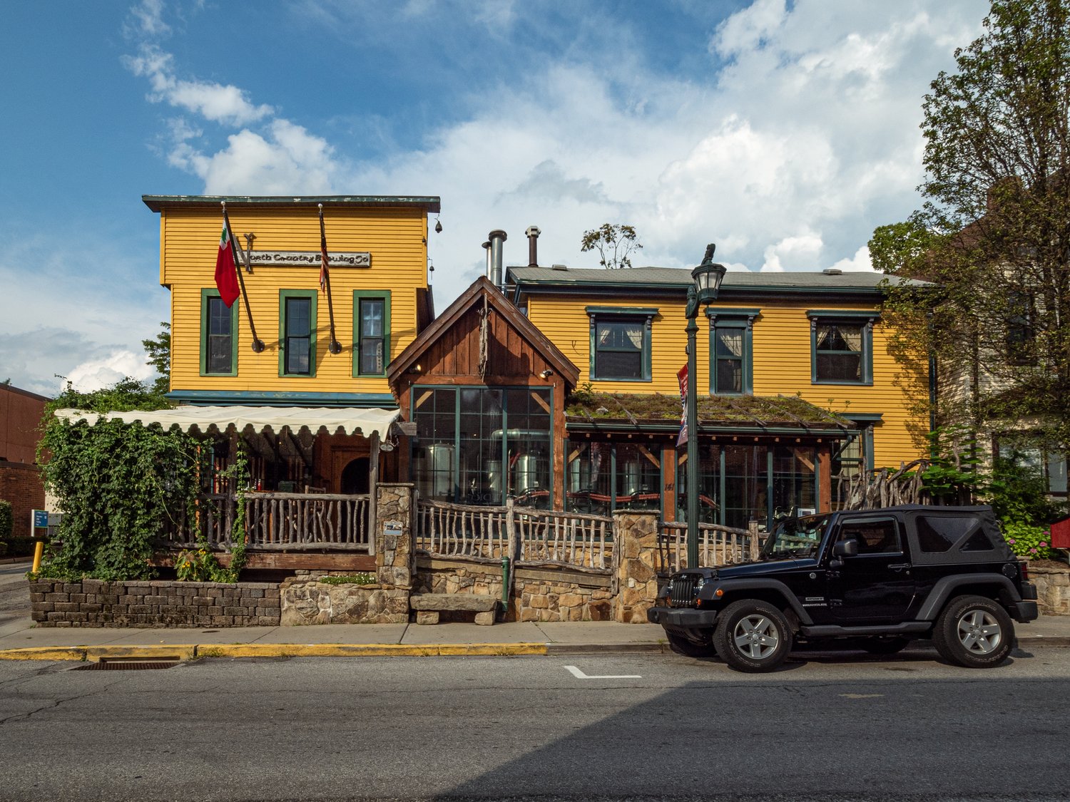 A yellow building with green shutters and a wooden covered front porch. A black jeep sits outside on the right in a street, parallel parking spot. 