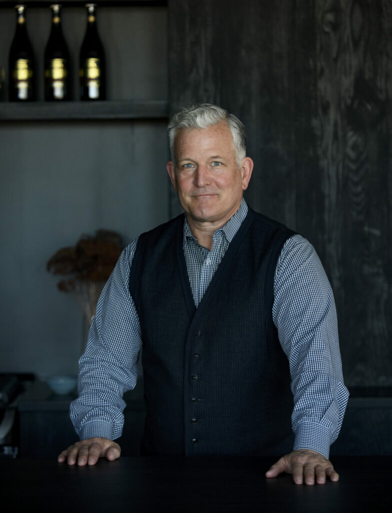Keith Recker stands at a black counter in a dark blue vest and long sleeve.