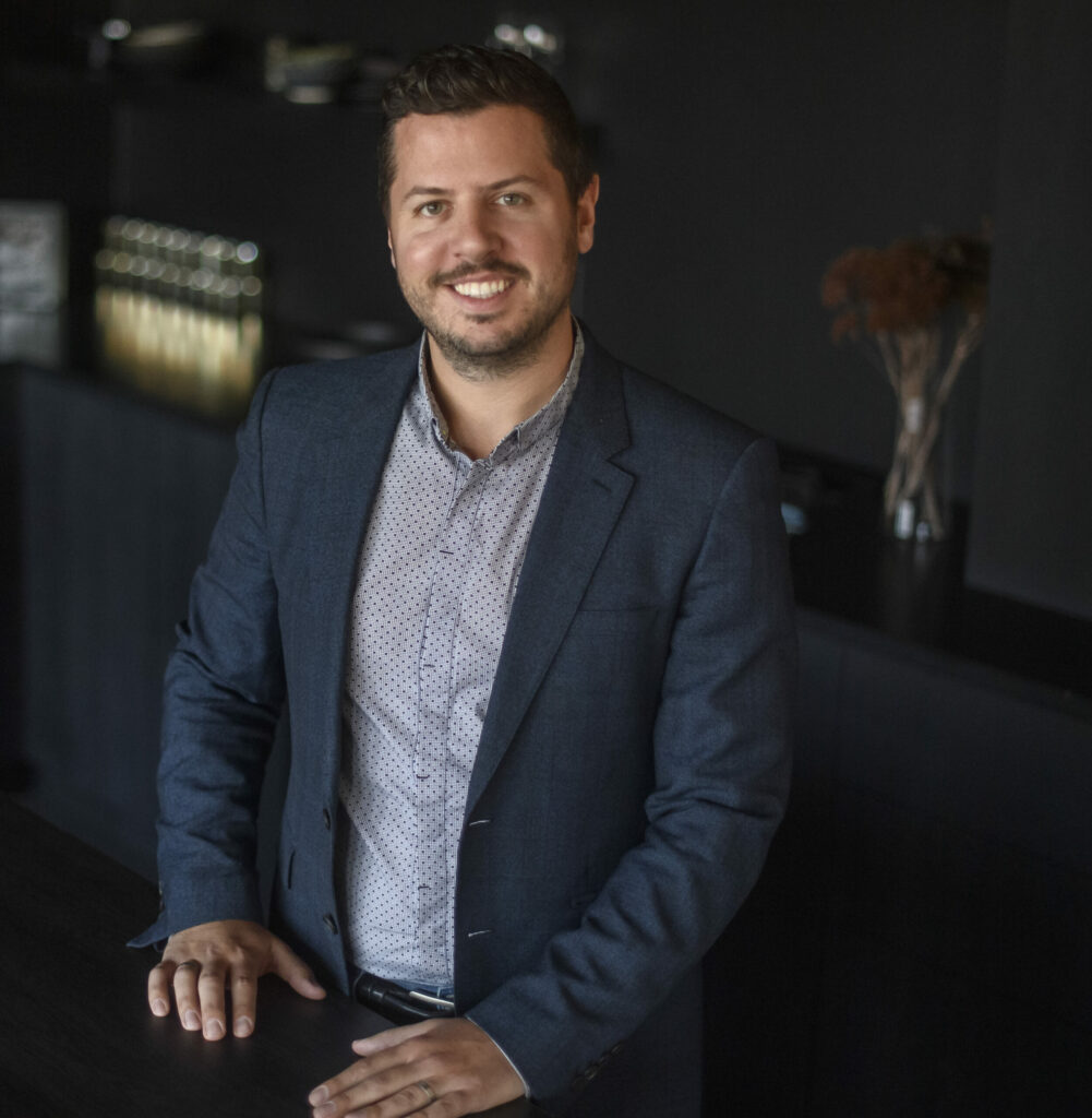 Justin Matase stands at a black counter in a blue button up and suit jacket.