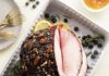 Lamb and Ham Recipes for Easter and Beyond An aerial view of a cooked ham with blueberry yuzu glaze sitting in a white roasting tray with blueberries, thyme, and lemon garnishing the meat.