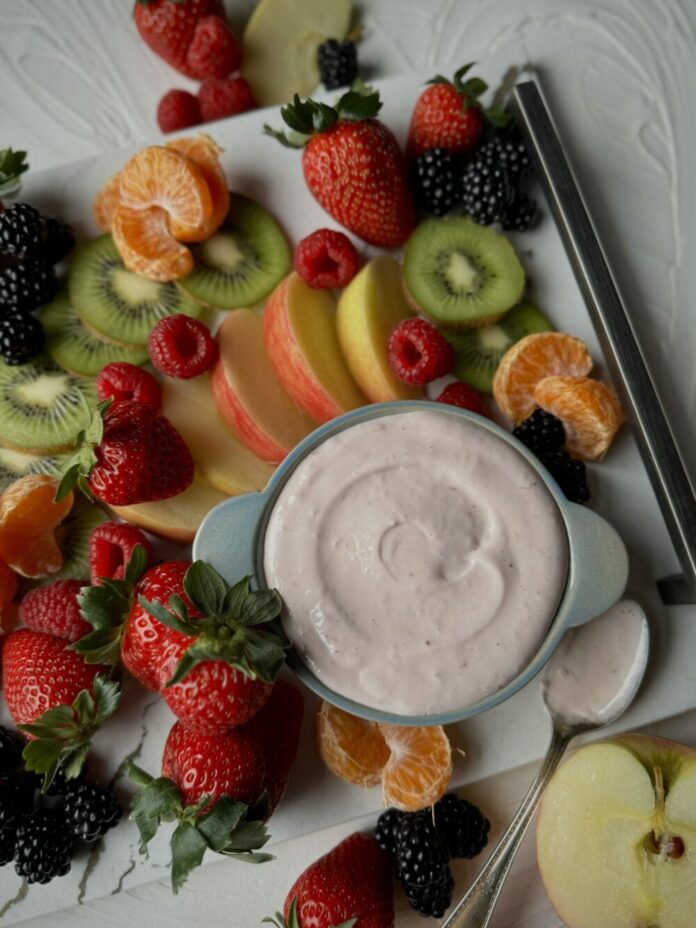 A bowl of easiest fruit dip in a light blue bowl on a white quartz surface surrounded by fresh strawberries, blackberries, raspberries, kiwi fruit, mandarin oranges, and apple slices.