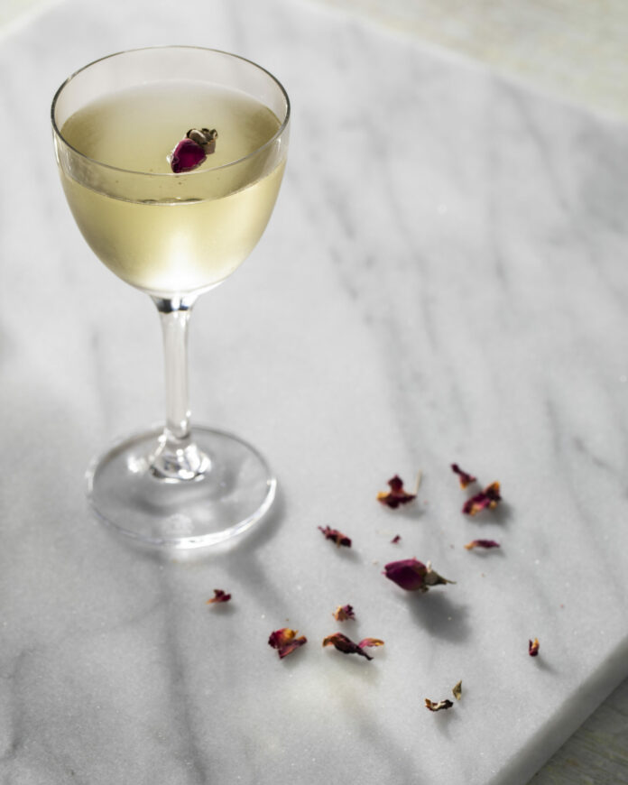Rose and Black Pepper Martini — Table A slightly yellow in color Rose and Black Pepper Martini in a martini glass sits on a white granite countertop. Rose petals are off to the side.