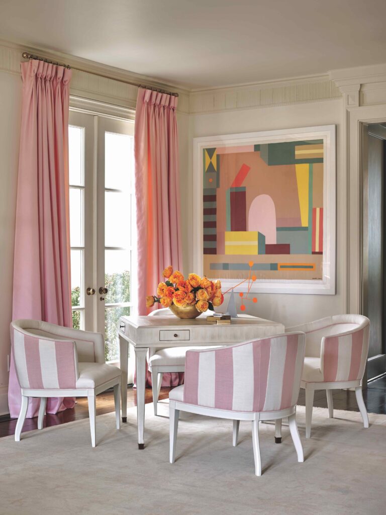 A photo of four pink striped chairs, a white desk, pink curtains and modern art. 