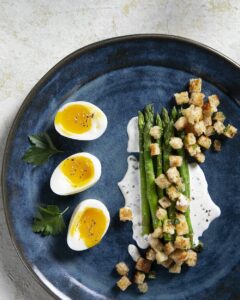 On a blue plate, three soft-boiled eggs sit on the left side while stalks of asparagus covered in Buttermilk Dressing and Sourdough Croutons sits to the right.