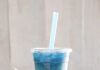 7 Pittsburgh Boba Tea Shops a look in photo with a rustic green table and a white paneled background of a to-go cup with boba tea that is a light tan color at the bottom with dark brown boba and a bright blue butterfly pea tea at the top, a blue straw