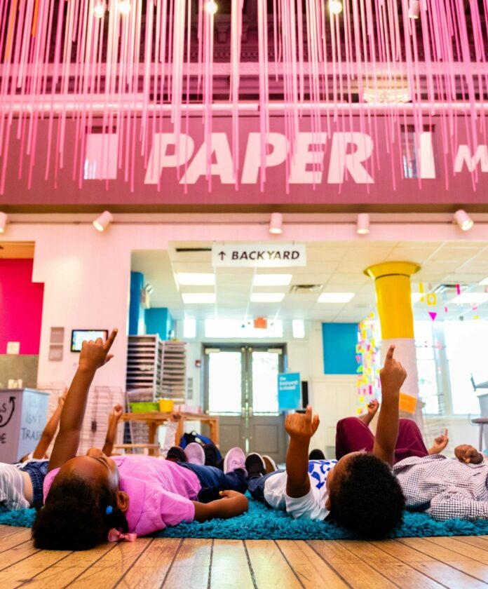 Kids lie on the floor pointing to the ceiling that is covered in pink string of dangling paper. They are at the Childrens Museum, Activities to do with Kids