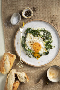 An aerial view of a dippy egg sitting on top of creamed spinach. a few pieces of sliced french bread sits off to the left of the plate.