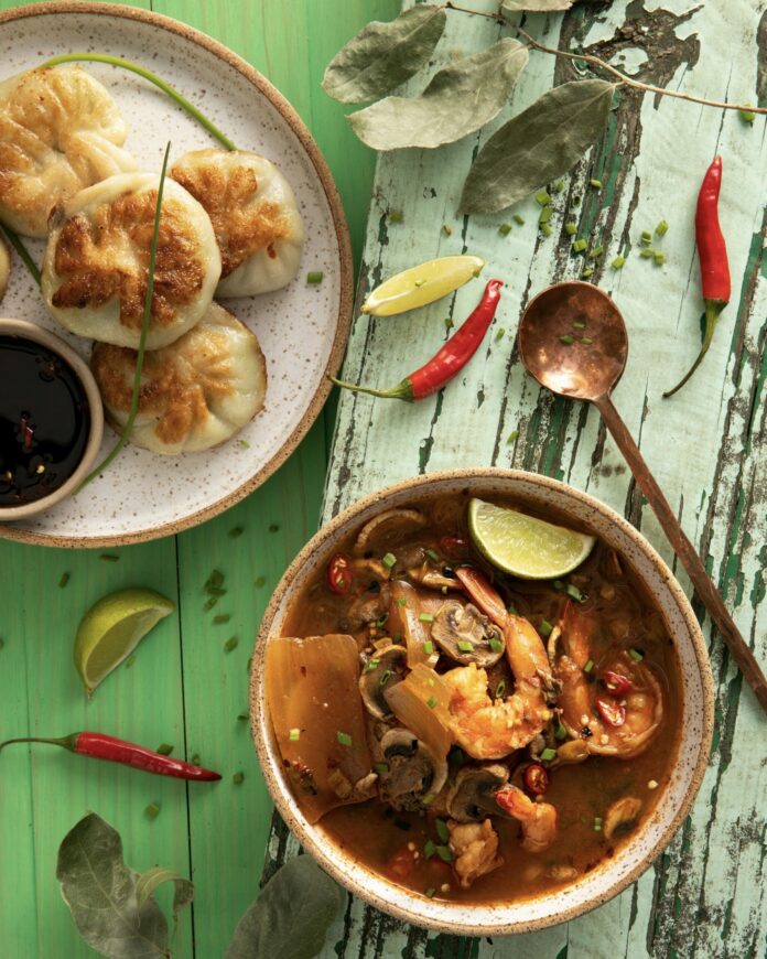 Chive Dumplings and Tom Yum Soup a tan plate with round dumplings and a bowl of red soup with shrimp on a green table