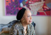 Jo-Anne Bates Goes Beyond Traditional Printmaking Jo-Ann Bates, a Black female artist, sits on the side of a chair and looks to the right. She wears a small black hat on her head and a zebra print button up.