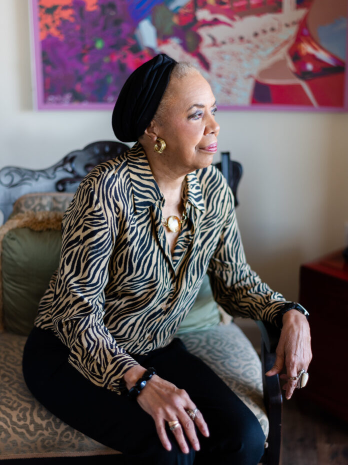 Jo-Ann Bates, a Black female artist, sits on the side of a chair and looks to the right. She wears a small black hat on her head and a zebra print button up.