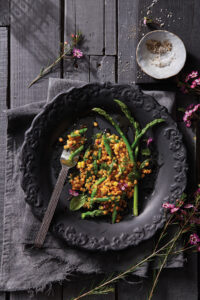 Orange lentil Asparagus Corn Salad sits on a black plate on top of a black wooden table.