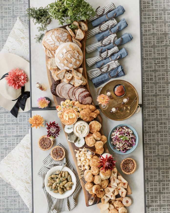 along board with meats and cheeses and other snack foods with a fancy hat to the left and blue and white cloth napkins rolled into horseshoe napkin rings on the right