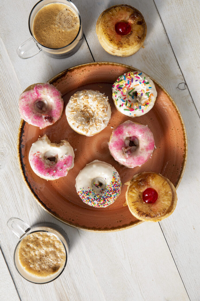 Seven multile colored donuts in Pittsburgh sits on an orange plate. One has a bite out of it. Two coffees sit around the plate, at the top and the bottom.