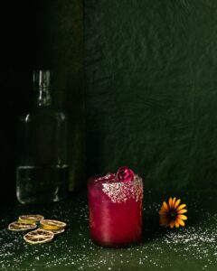 A dark-pinkish red prickly pear margarita sits in a salt-rimmed glass in front of a green background.