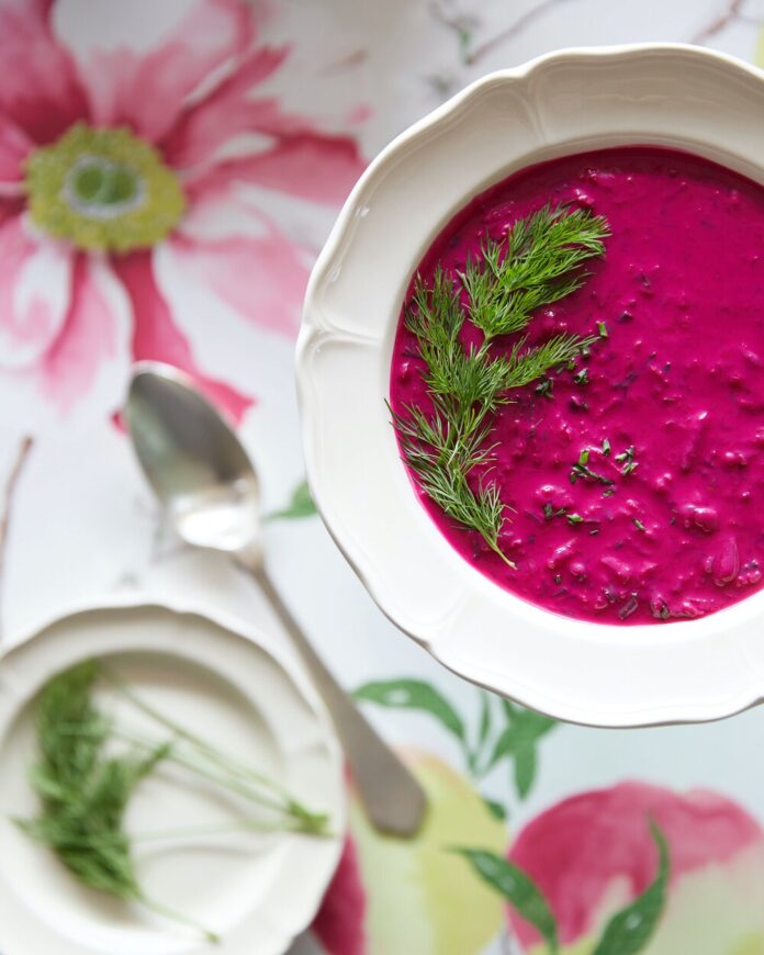 Chlodnik soup in a bowl alongside a blurred background with a spoon and flowered sheet on a table.