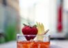 Strawberry Negroni Bianco A Strawberry Negroni Bianco sits in a rocks glass, garnished with a strawberry and twist of a peel of lemon. A hand holds the glass on the right.