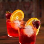 Two Strawberry Rhubarb Negronis sit in short glasses, one slightly in front of the other. Deep red in color, they are garnished with a strawberry and orange slice.