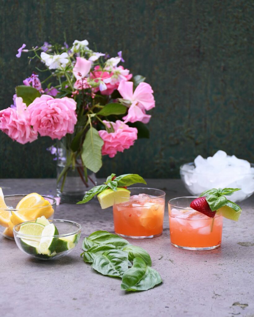 Next to a large vase of flowers sits two strawberry farm cocktails in short rocks glasses, garnished with basil.