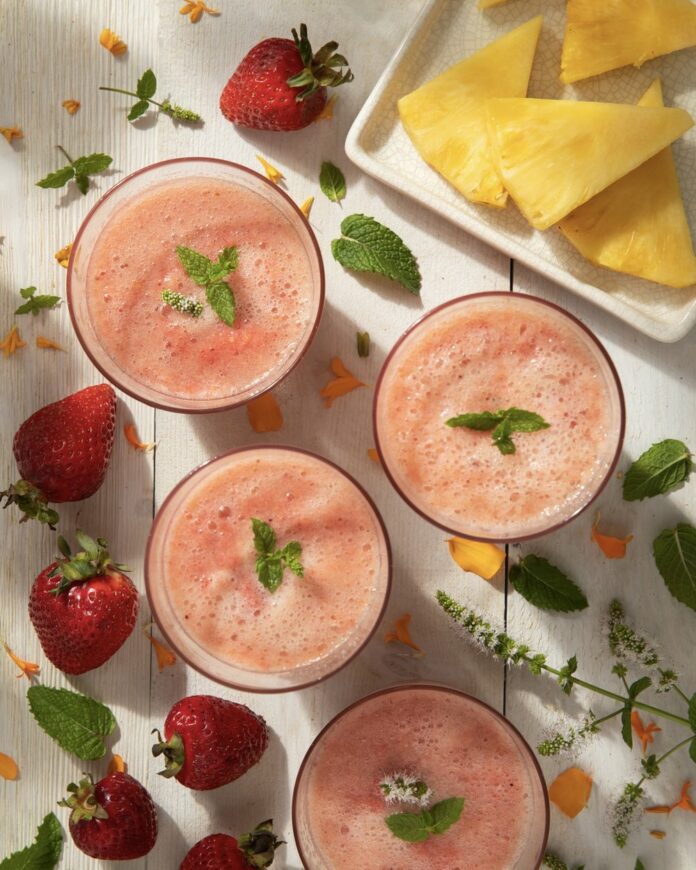 Three glasses of strawberry pineapple agua fresca sit in glasses on a white picnic table surrounded by strawberries and slices of pineapple.