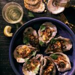 7 roasted oyster with corn and herb garnish on a blue plate and 2 oyster shells and a beer sitting on a dark colored surface. seafood dishes