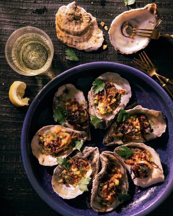 7 roasted oyster with corn and herb garnish on a blue plate and 2 oyster shells and a beer sitting on a dark colored surface. seafood dishes