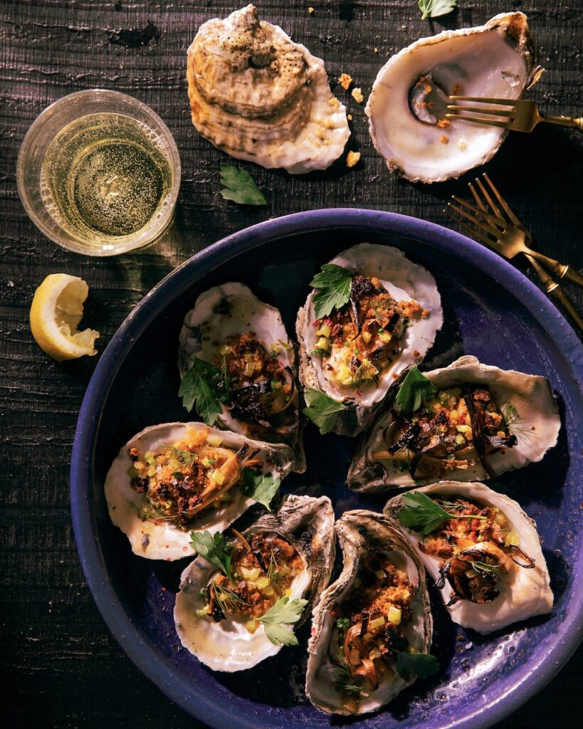 7 roasted oyster with corn and herb garnish on a blue plate and 2 oyster shells and a beer sitting on a dark colored surface. seafood dishes