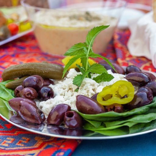 an appetizer plate of olives, pickles and cheese on a table covered with a colorful tablecloth of red orange and blue