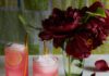Hibiscus‑Mint Spritz Two skinny glasses of our Hibiscus and Mint Spritz cocktail sit to the left next two a vase of full, blooming, dark red flowers.