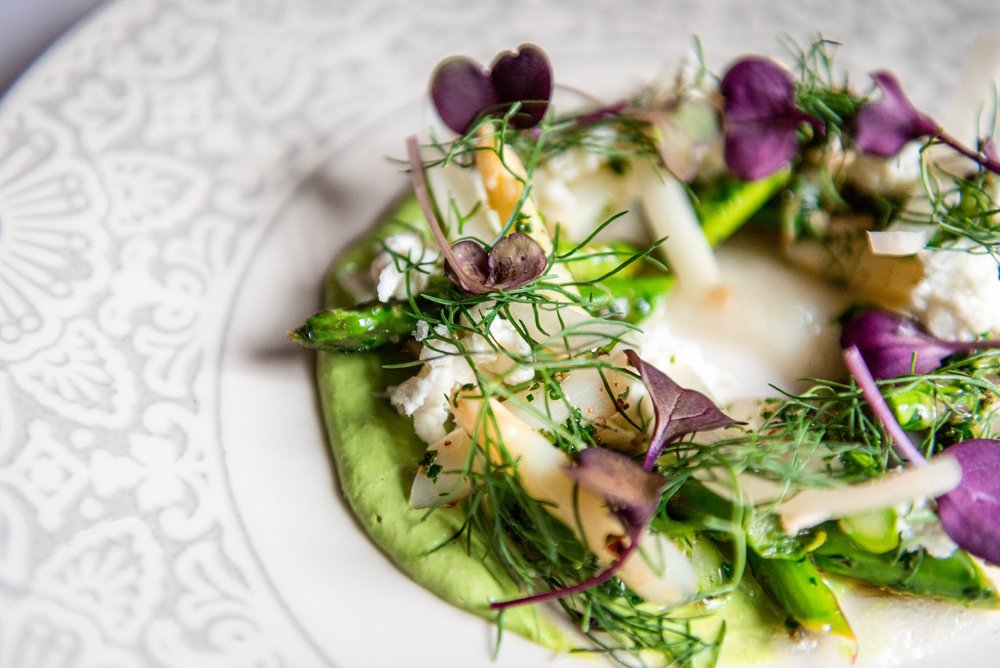 a white plate with a vibrant green vegetable puree, white and green asparagus, dill and purple edible flowers