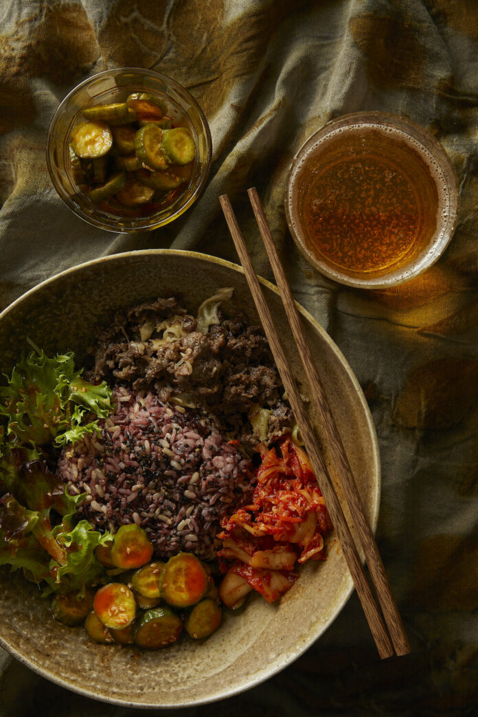 Cucumber Kimchi in a large bowl with two sticks on top of it. On the side, lies a drink and a small bowl of sliced cucumbers