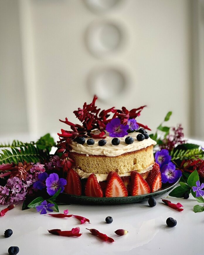 Strawberries, berries, and flowers adorn a ginger cake with the sides non-iced, blue, pink, and purples color the cake.