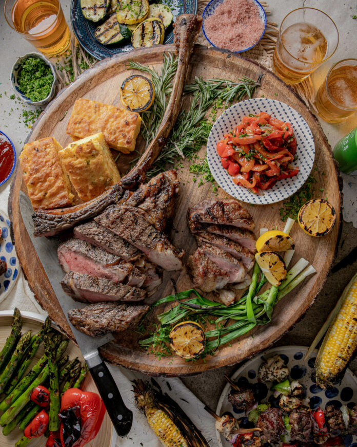 a round wooden board with a cut tomahawk steak, corn bread, an individual steak, roasted red peppers, grilled green onion, and grilled yellow lemons with other vegetables and accruements including grilled corn on the cob and glasses of beer.