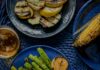How to Grill Corn and Fresh Vegetables a blue plate on a blue woven placemat with grilled asparagus and grilled lemons with a separate plat of grilled squash and a separate plate of grilled corn on the cob, and a glass of beer