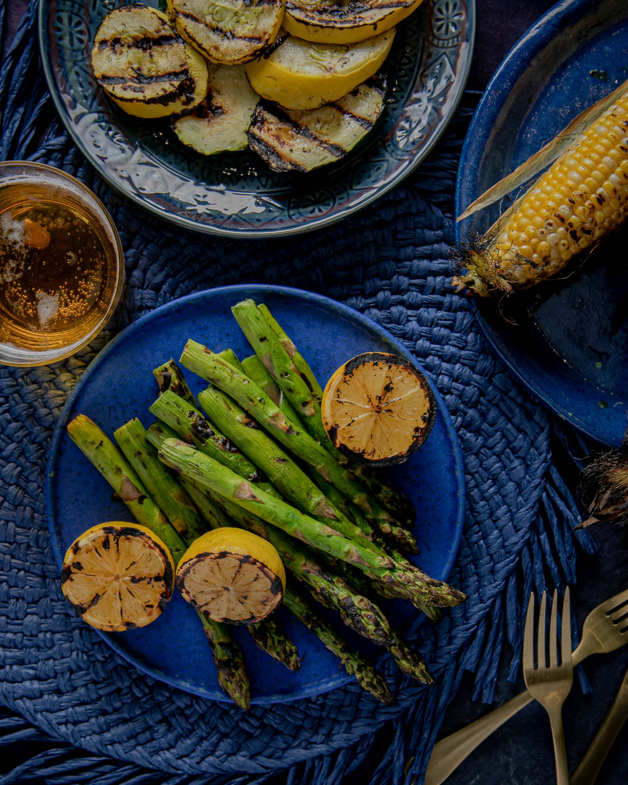 a blue plate on a blue woven placemat with grilled asparagus and grilled lemons with a separate plat of grilled squash and a separate plate of grilled corn on the cob, and a glass of beer