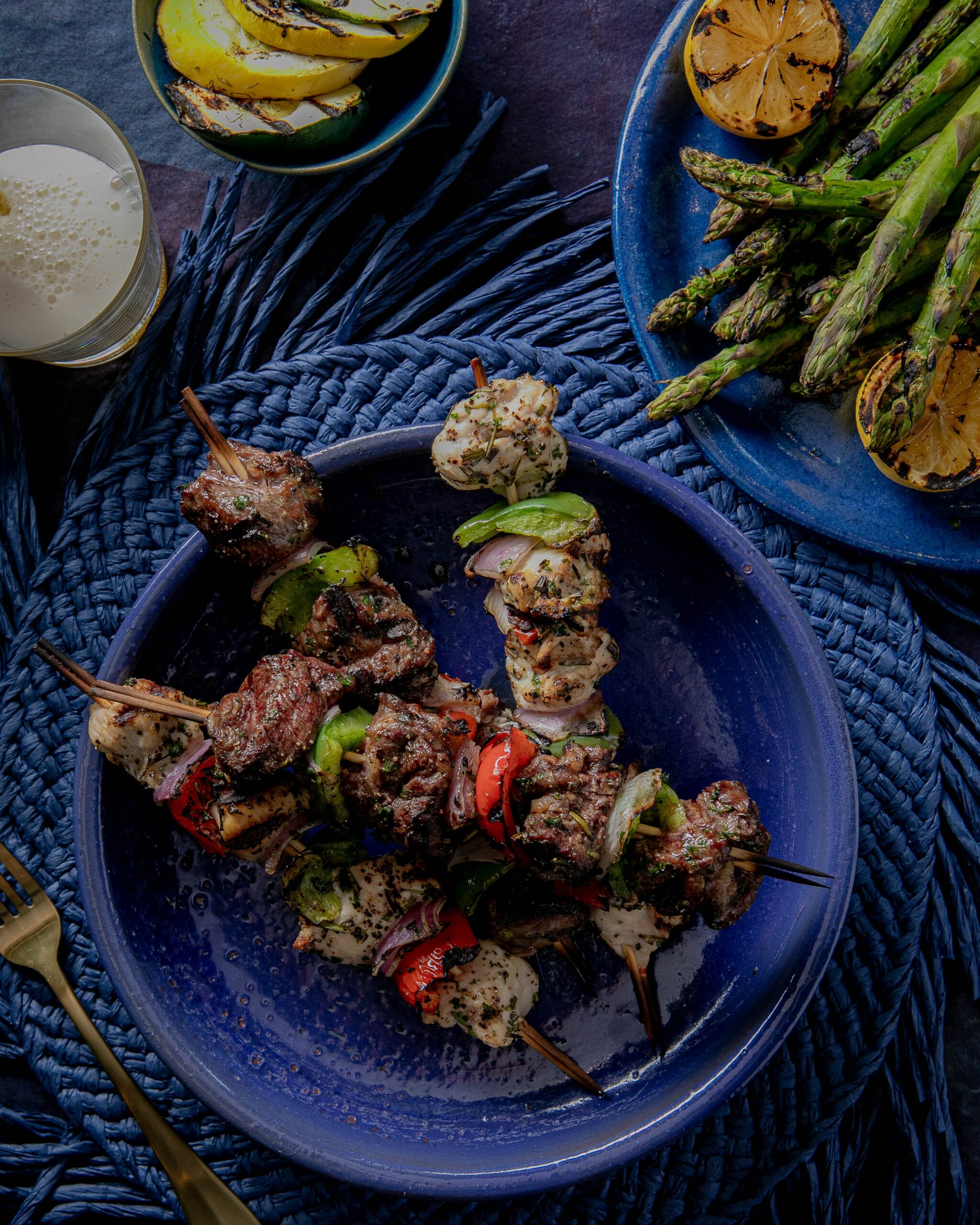 Skewers with beef, chicken, and vegetables on a blue plate and blue straw placemat with a glass of beer and a separate plate of grilled vegetables