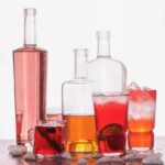 A variety of red bottles for a summer punch sit on a table with various mixed red and orange cocktails in glasses in front of the bottles.