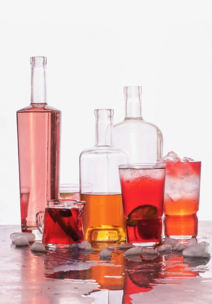 A variety of red bottles for a summer punch sit on a table with various mixed red and orange cocktails in glasses in front of the bottles.
