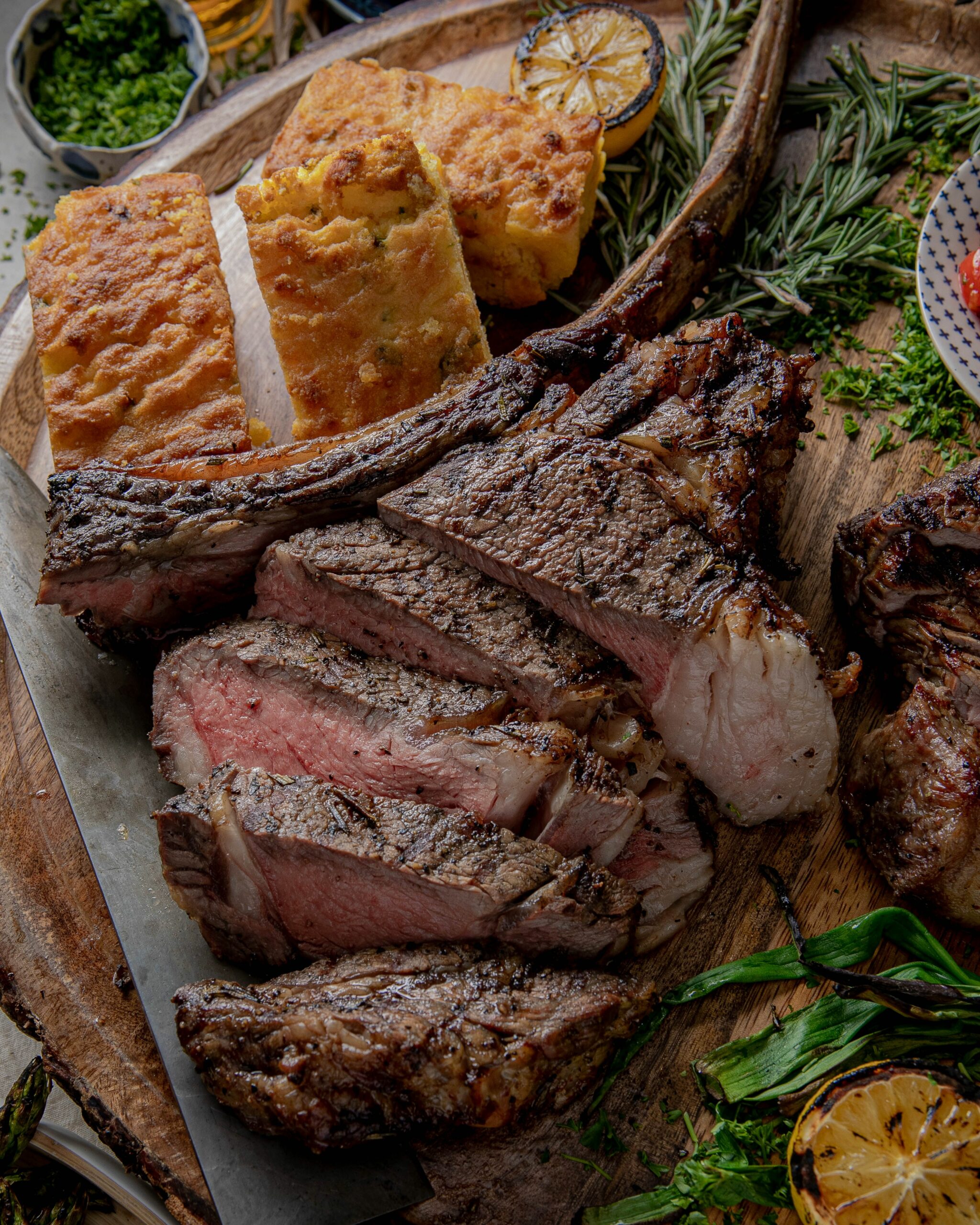 a medium rare tomahawk steak cut into several pieces and placed on a board with corn bread, grilled green onions and grilled lemons