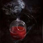 a deep red cocktail in a hanging glass with a small bird hole type opening with a dark background and smoke