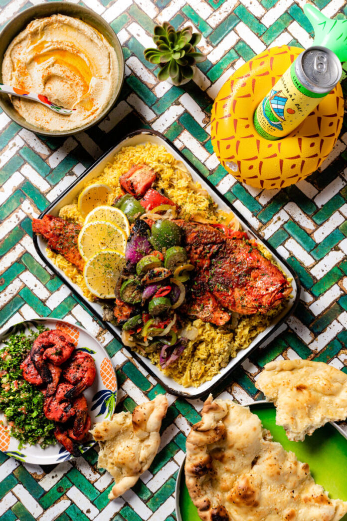 Hummus, Grilled Red Snapper & Vegetables over Biryani, garlic naan, tandoori shrimp and tabbouleh from Salem’s Market & Grill and Locally made Personal Day Hard Seltzer from Maggie's Rum Farm against green and white tile for BBQ season