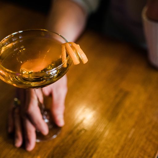 a cocktail in a coupe glass with an orange garnish and a man's hand around the base of the glass resting on a wooden table