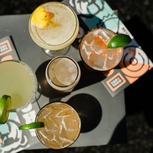 An overhead shot of 5 cocktails. The one at the top has a yellow rubber ducky, the middle one is a smaller glass and the 3 outer drinks are garnished with green limes