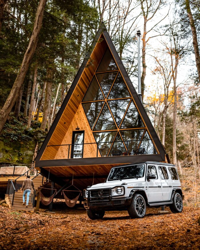 An A frame cabin with big glass windows in the woods. In front sits a grey/white Jeep to the right of the image. Long Weekend idea for Pittsburgh Travel