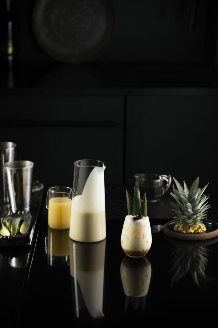 A piña colada mocktail, white in color, sits in a stemless wine glass next to a glass pitcher of the mixed drink.