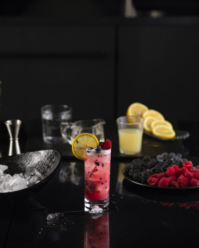 Wildberry Sparkler: A Refreshing Mocktail red in color, sits in a glass filled with berries and garnished with lemon.