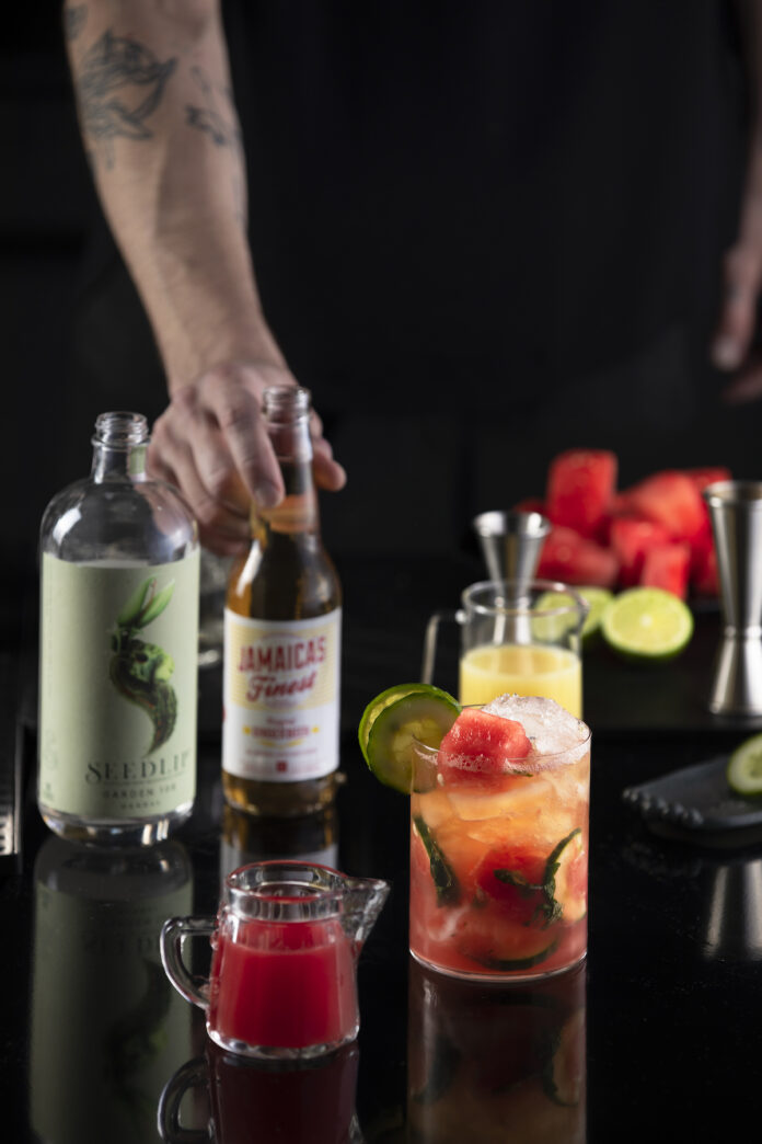 A white man's hand hold a bottle of ginger beer as it sits on a black table. Next to the soda sits a mojito mocktail orange in color.